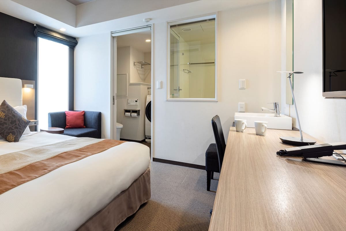 Quarto de hotel com uma cama grande, uma escrivaninha, uma cadeira, área de chuveiro e vista para a cidade.