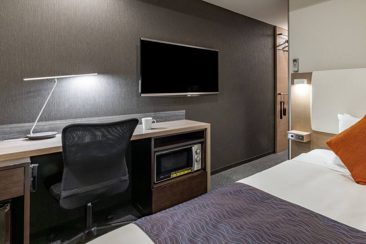 Quarto de hotel com uma cama, escrivaninha com computador, micro-ondas, televisão e mesa de cabeceira com abajur.