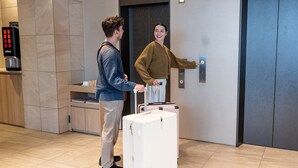 A man and a woman standing in front of an elevator in a modern building with a coffee machine and a wooden counter in the background. - Tokyu Stay Shimbashi (Ginza area) (Tokyo)