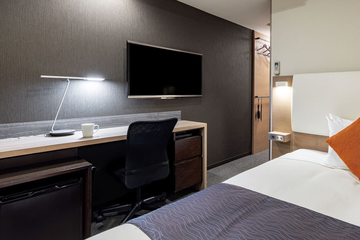 Quarto de hotel com cama, escrivaninha, cadeira, televisão e mesa de cabeceira.