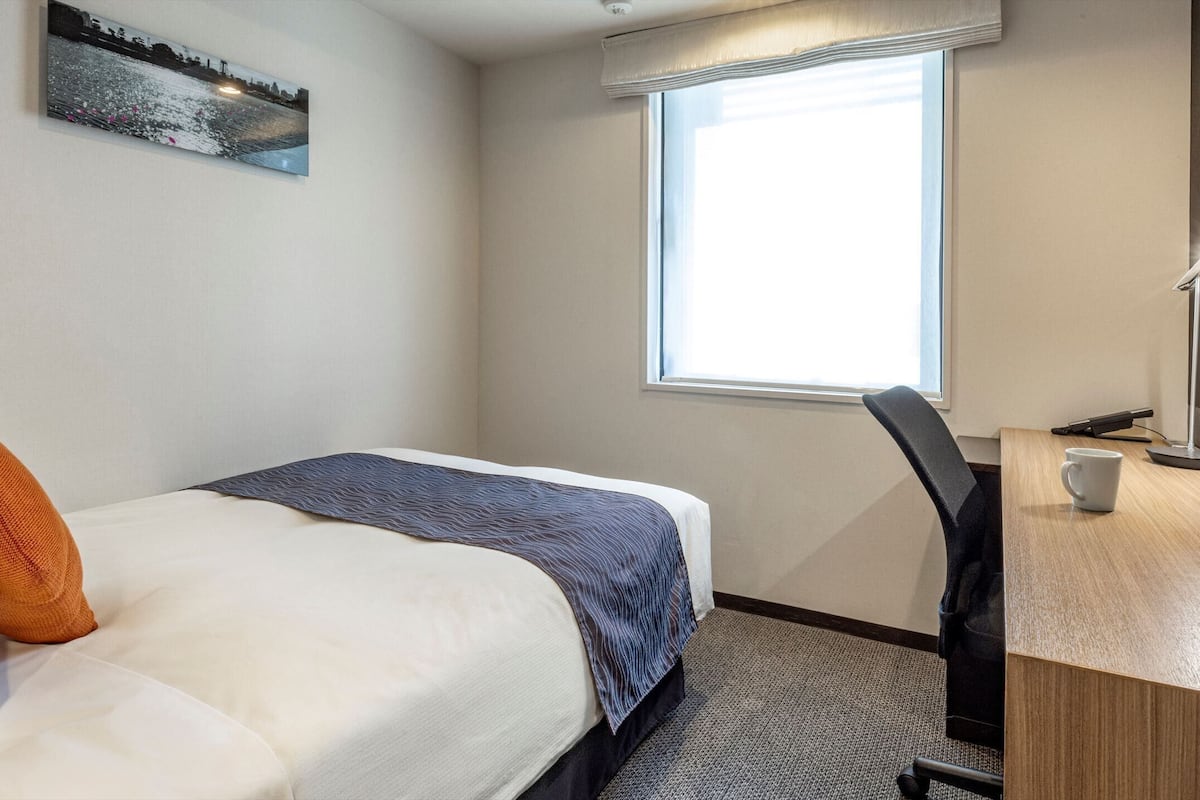 Quarto de hotel com cama, escrivaninha, cadeira, janela e um quadro na parede.