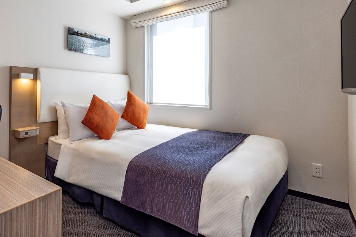 Quarto de hotel com cama, escrivaninha, cadeira, luminária, quadro na parede e janela.
