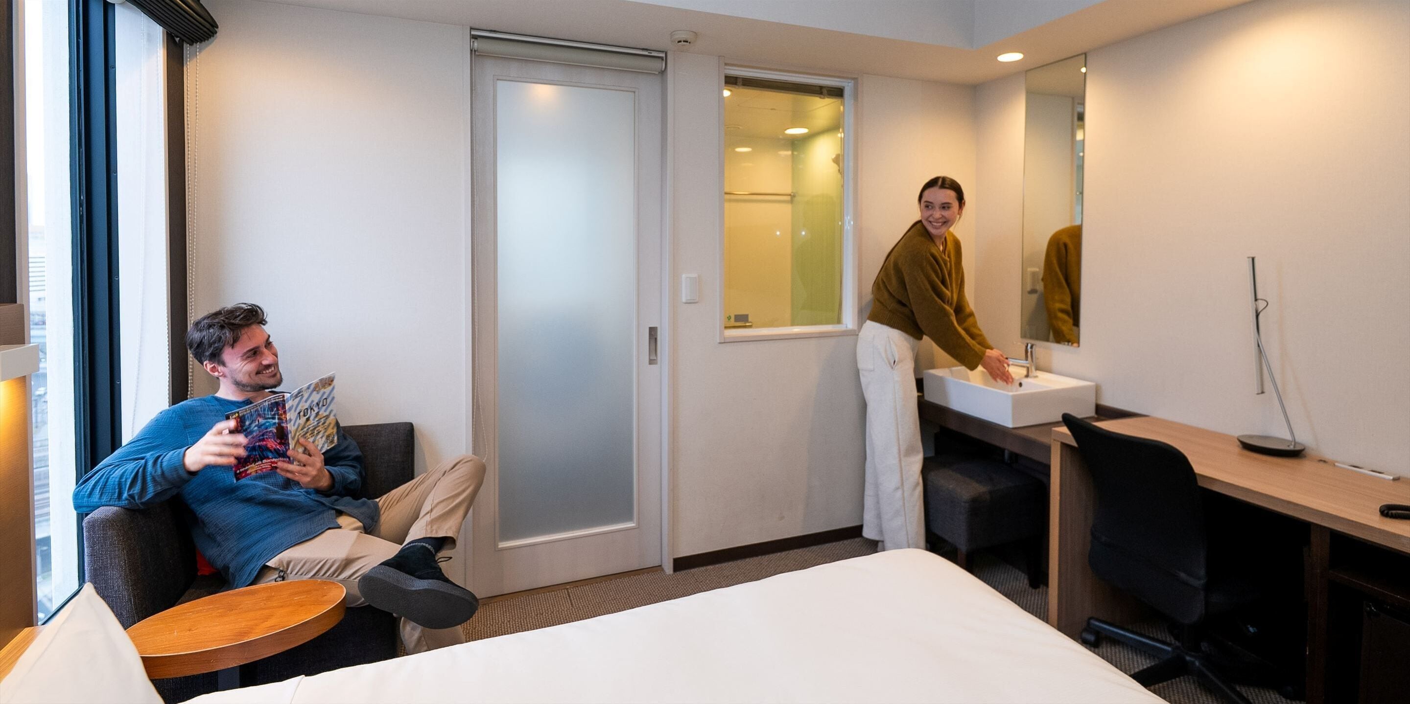 A hotel room with a bed, a sitting area with a table, a desk with a chair, a bathroom with a mirror, and a window.