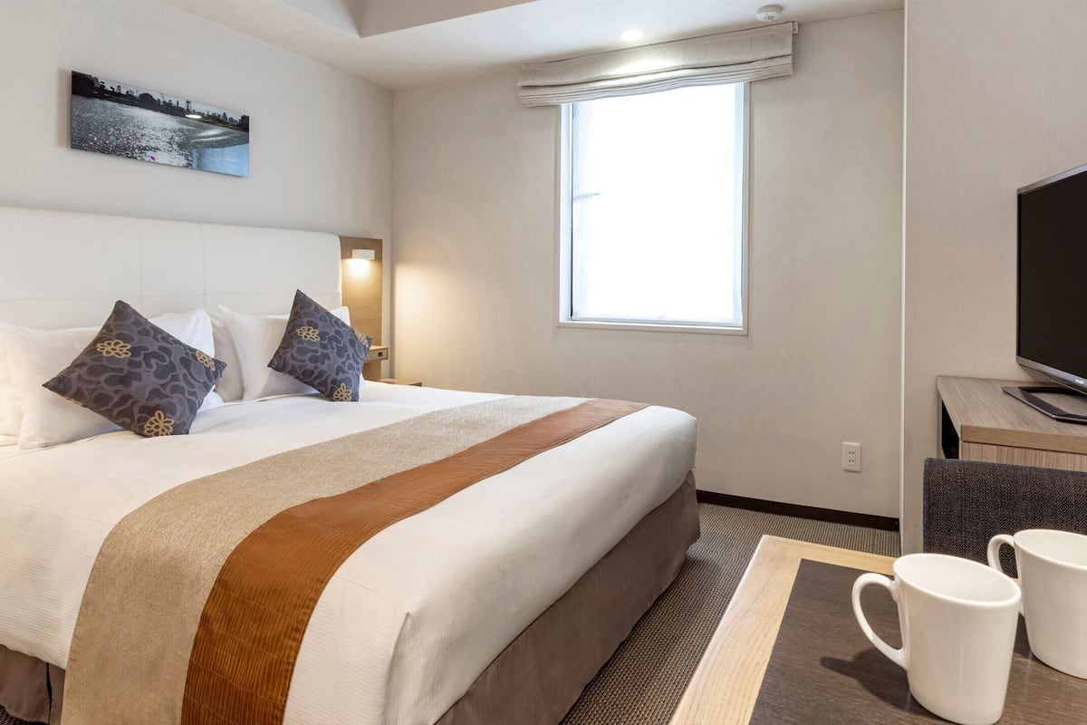Quarto de hotel com uma cama grande, uma TV de tela plana e duas canecas brancas sobre uma mesa.