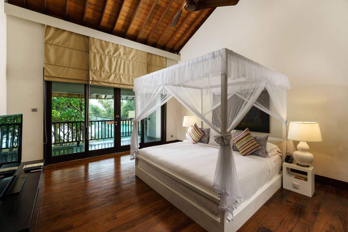 A four-poster bed with a canopy, bedside lamps, and a view of greenery through large windows.