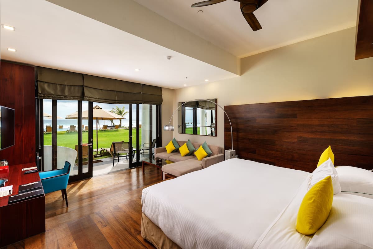 A hotel room with a large bed, a desk, and a view of the beach through the glass doors.