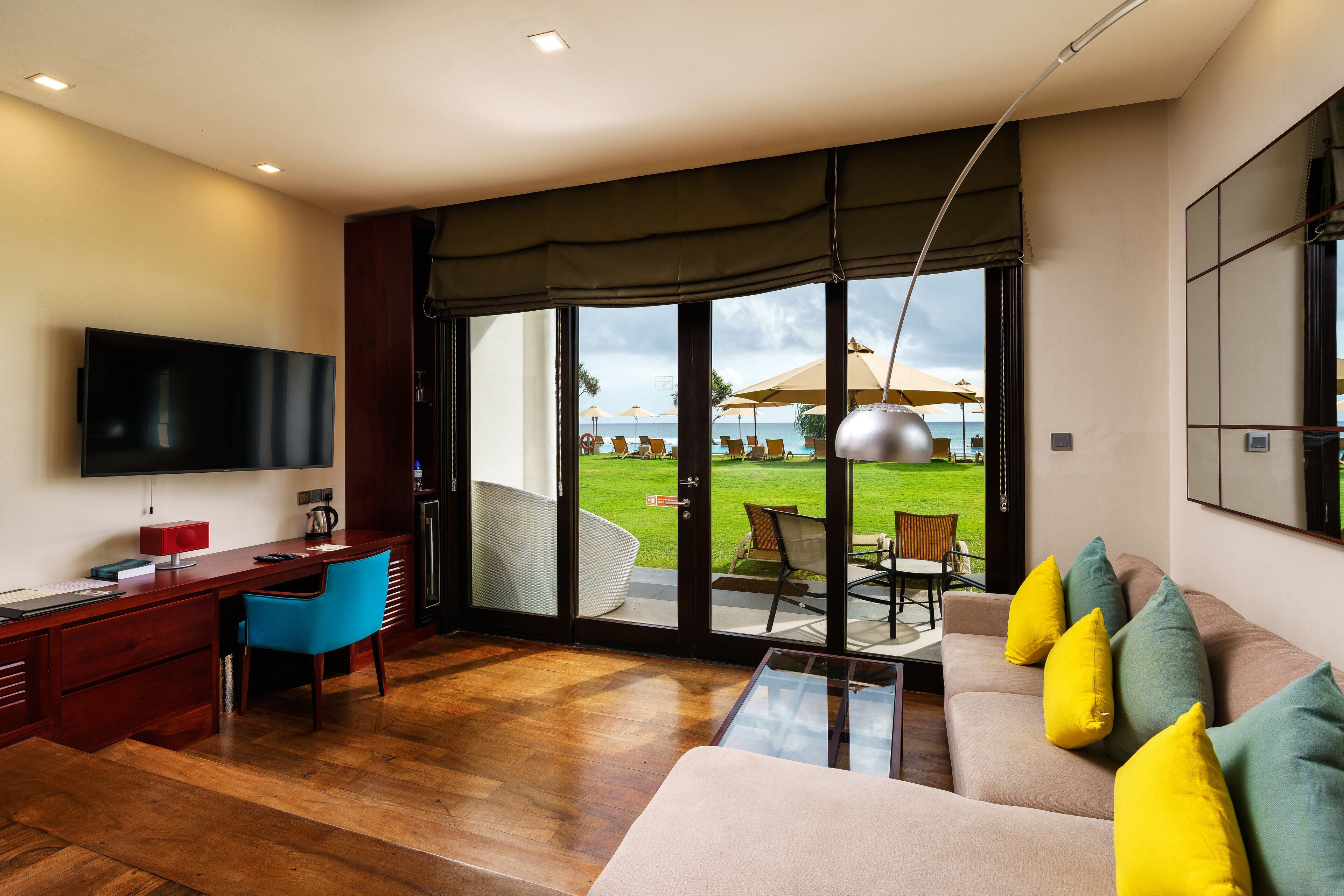 A modern living room with a wooden floor, a sofa with yellow cushions, a blue chair, a desk with a TV, and a large sliding glass door leading to an outdoor area with umbrellas.