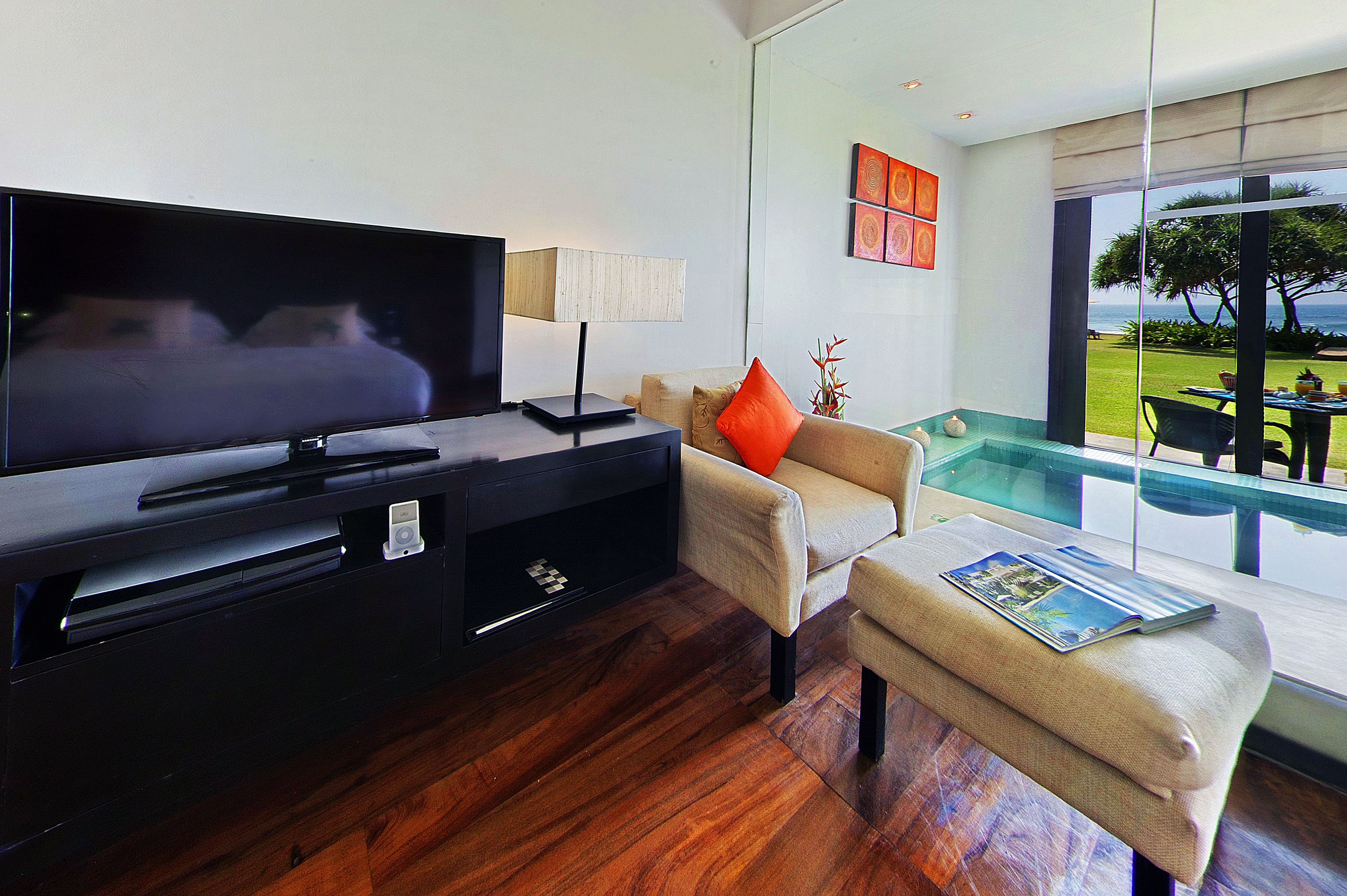 A modern living room with a flat-screen TV, a sofa with an orange pillow, a small table with a magazine, and a glass-enclosed pool area with a view of the ocean.