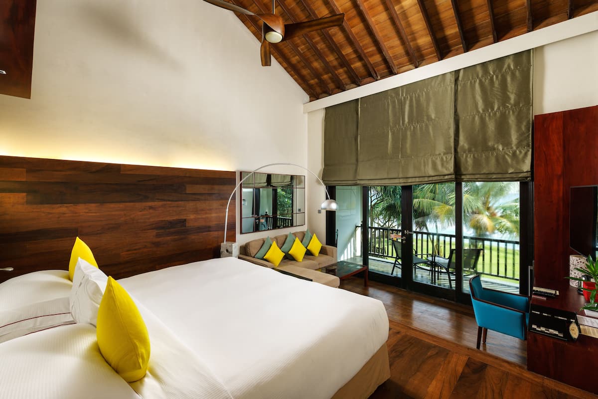 A modern hotel room with a large bed, a wooden ceiling, a balcony with a view of palm trees, and a desk with a chair.