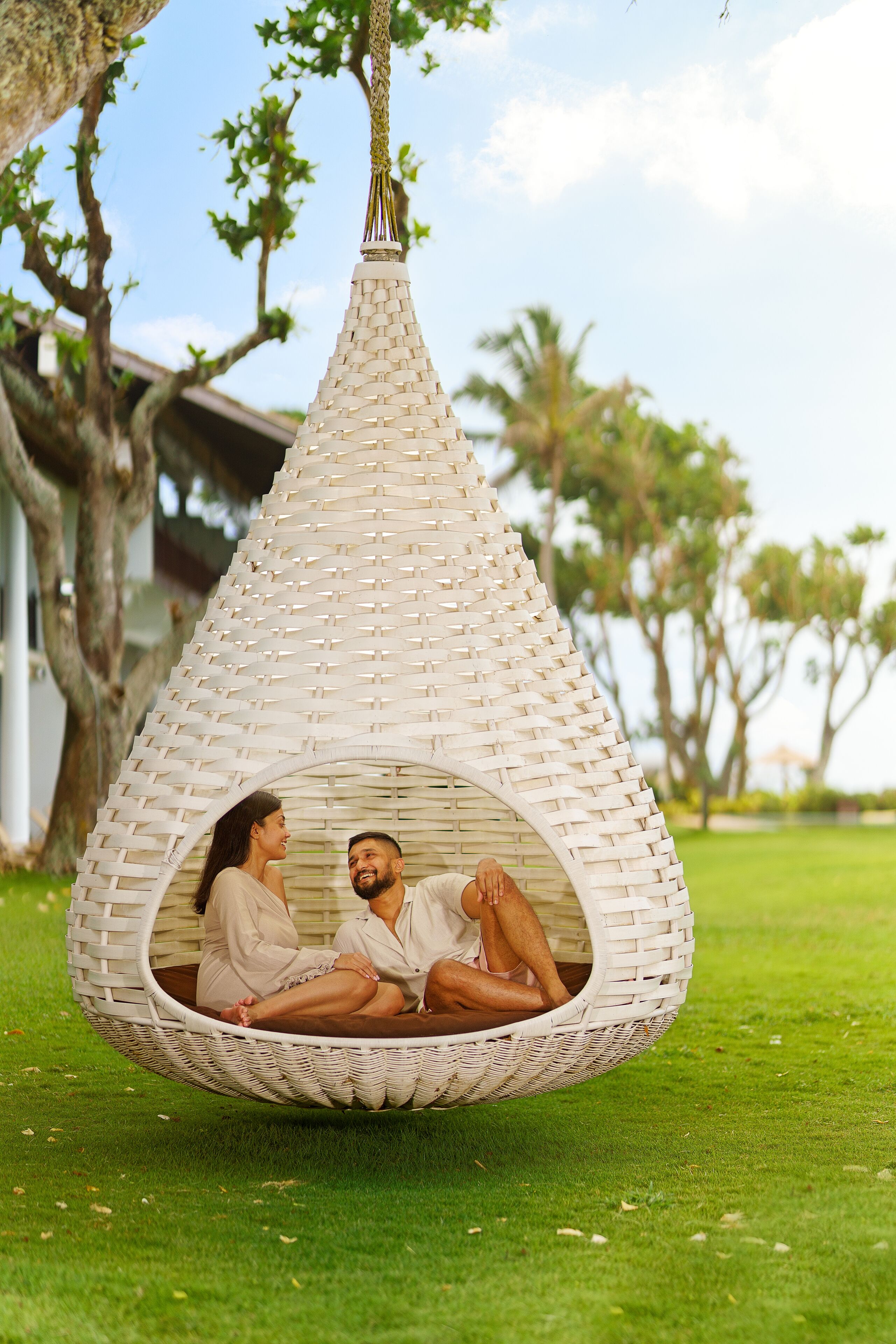 A couple sitting in a hanging wicker chair on a grassy lawn with trees in the background.