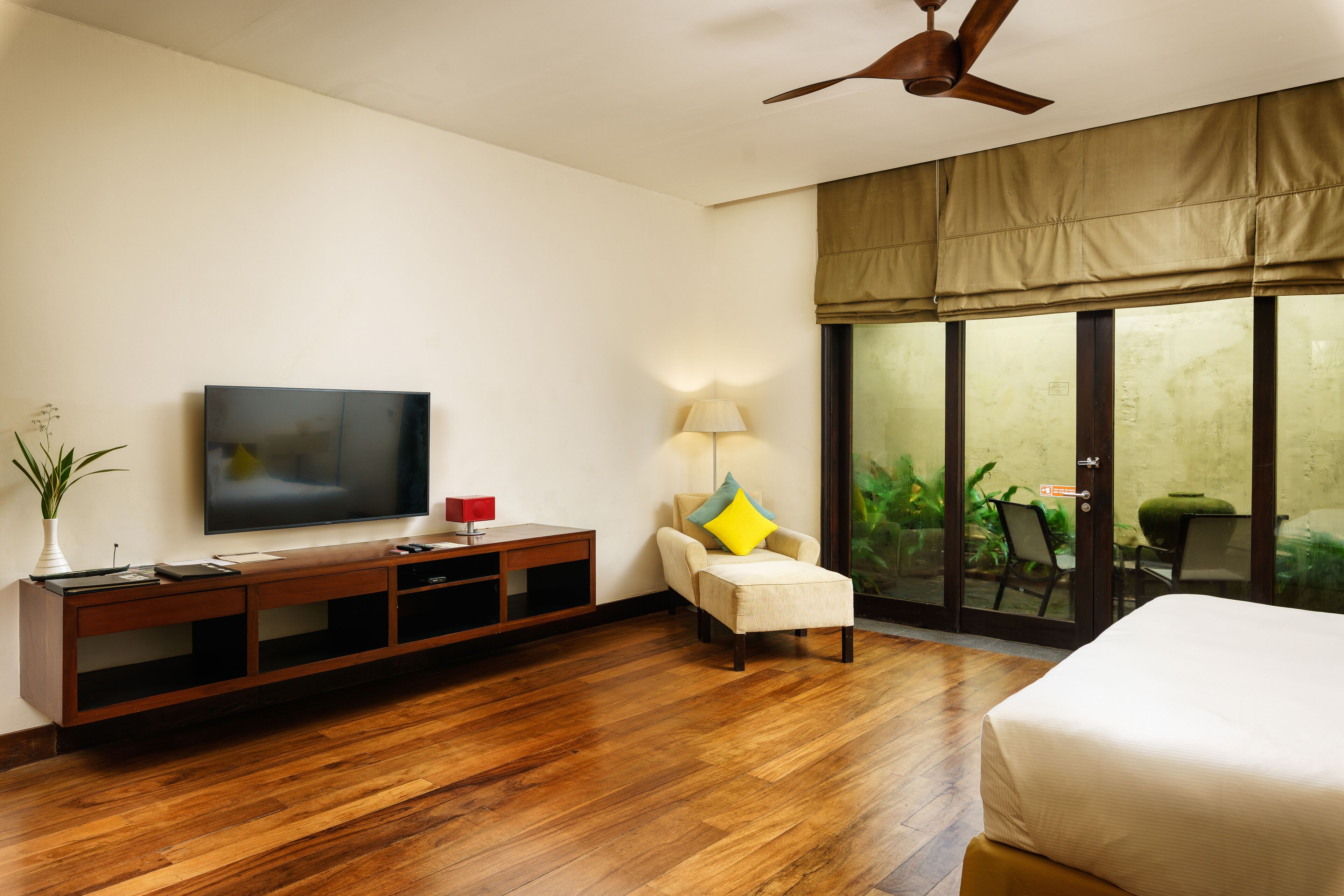 Habitación de hotel con suelo de madera, un televisor de pantalla plana sobre un mueble de madera, una silla con almohada amarilla, una cama y una puerta de vidrio corredera que da a un área exterior.