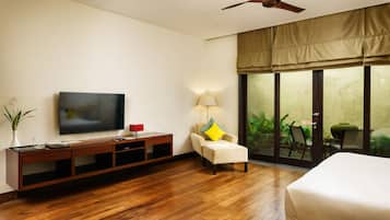 A hotel room with a wooden floor, a flat-screen TV on a wooden console, a chair with a yellow pillow, a bed, and a sliding glass door leading to an outdoor area.