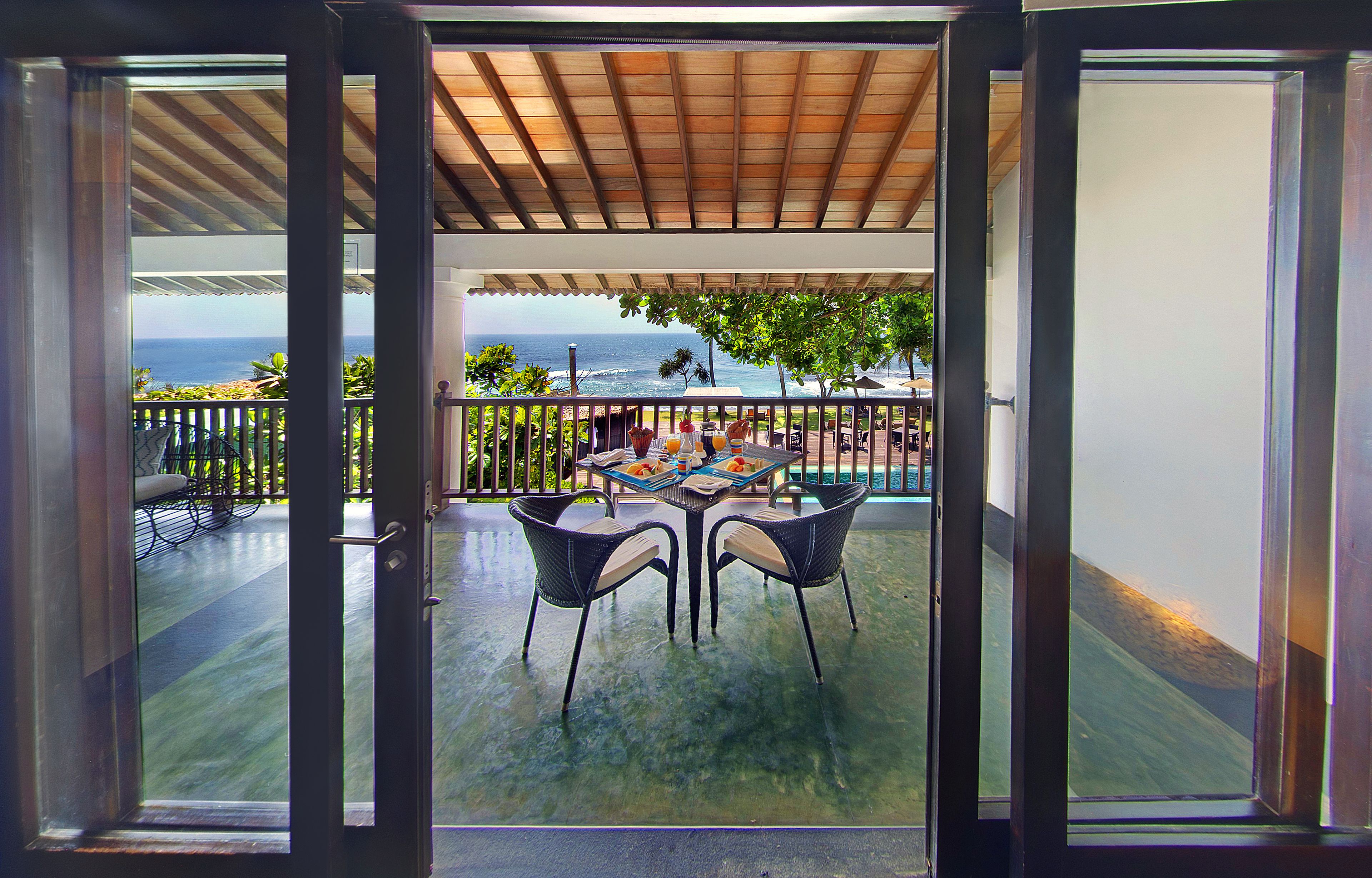 An open glass door leading to a covered outdoor area with a table and chairs, a view of the ocean, and a wooden ceiling.