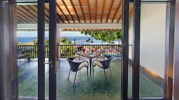 An open glass door leading to a covered outdoor area with a table and chairs, a view of the ocean, and a wooden ceiling.