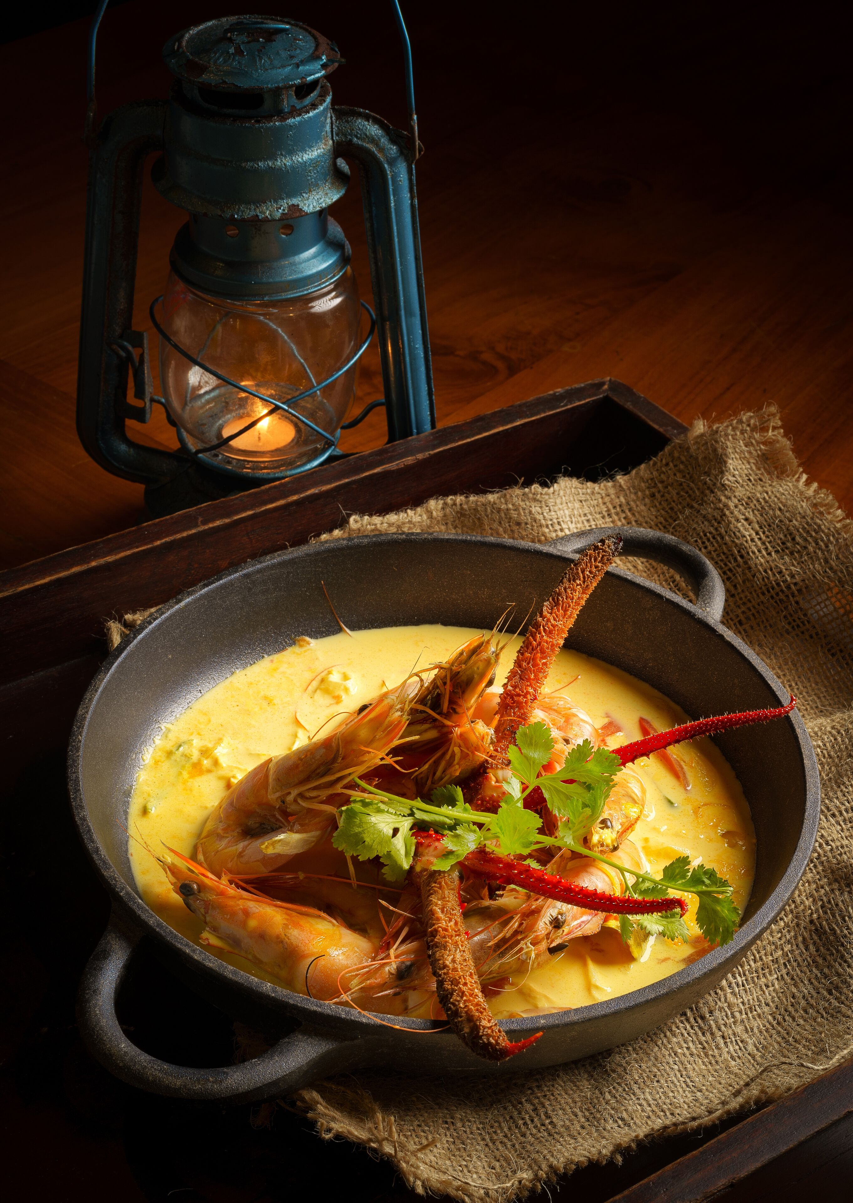 A pan of seafood soup with shrimp and a red chili, placed on a wooden table with a burlap cloth and a lit lantern in the background.
