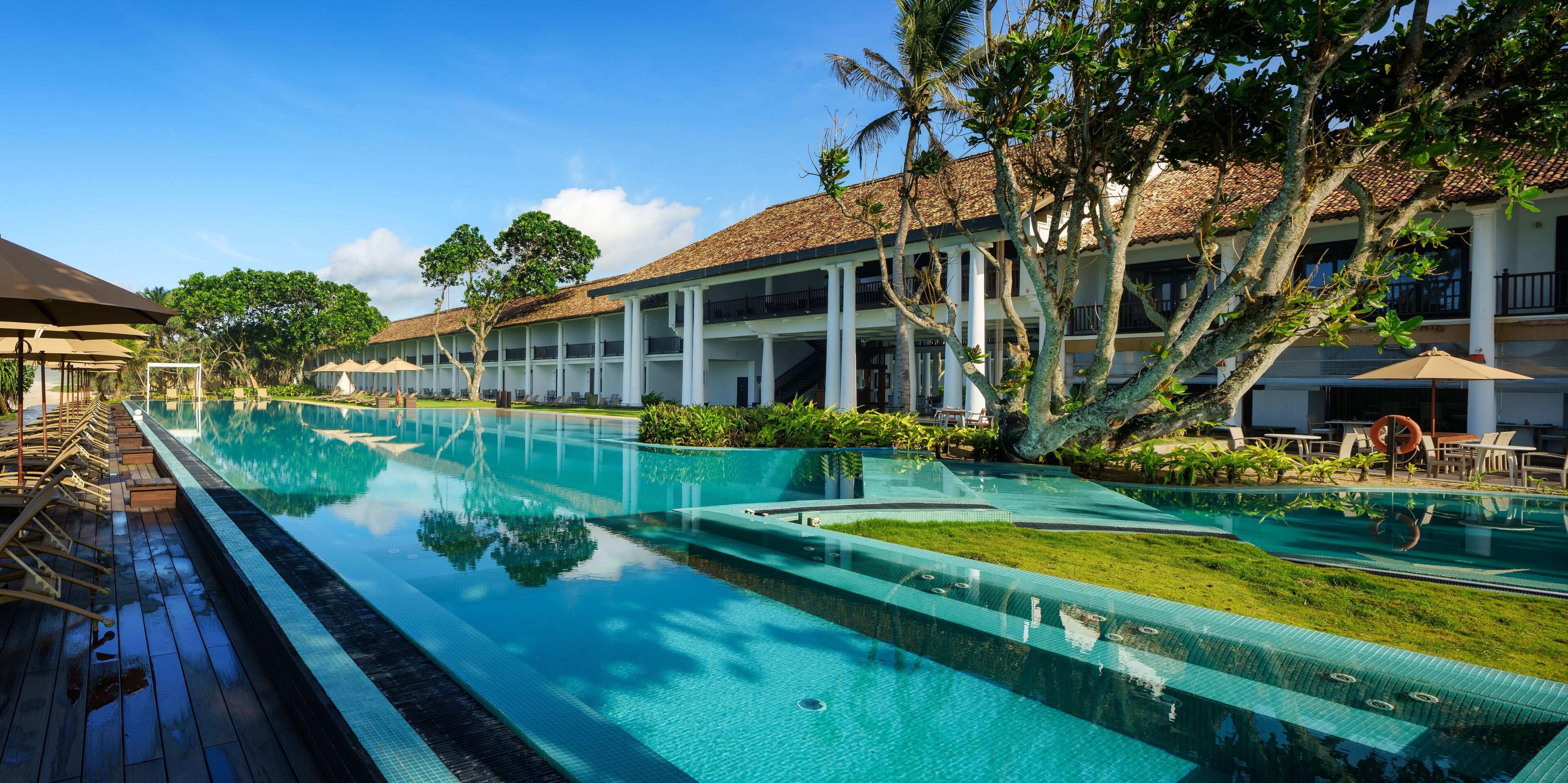 A resort pool area with clear blue water, wooden deck, and white buildings with balconies.