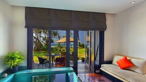A modern living room with a glass-enclosed pool, a sofa with a red pillow, and a view of a garden and beach. - The Fortress Resort & Spa Galle (Koggala)