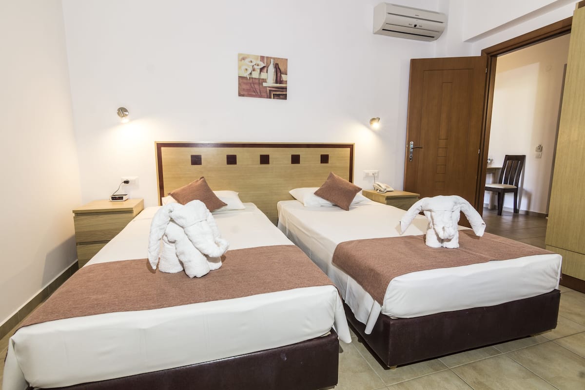 Habitación de hotel con dos camas, cada una con cabecero marrón, ropa blanca y un albornoz blanco sobre la cama. Hay un cuadro en la pared encima de las camas y un equipo de aire acondicionado en la pared.