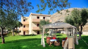A grassy outdoor area with a covered seating area, a building in the background, and trees. - Lindos Blue Sky (Rhodes)