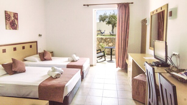 A hotel room with two beds, a desk with a computer, a TV, and a view of an outdoor patio with a hammock and a table. - Lindos Blue Sky (Rhodes)