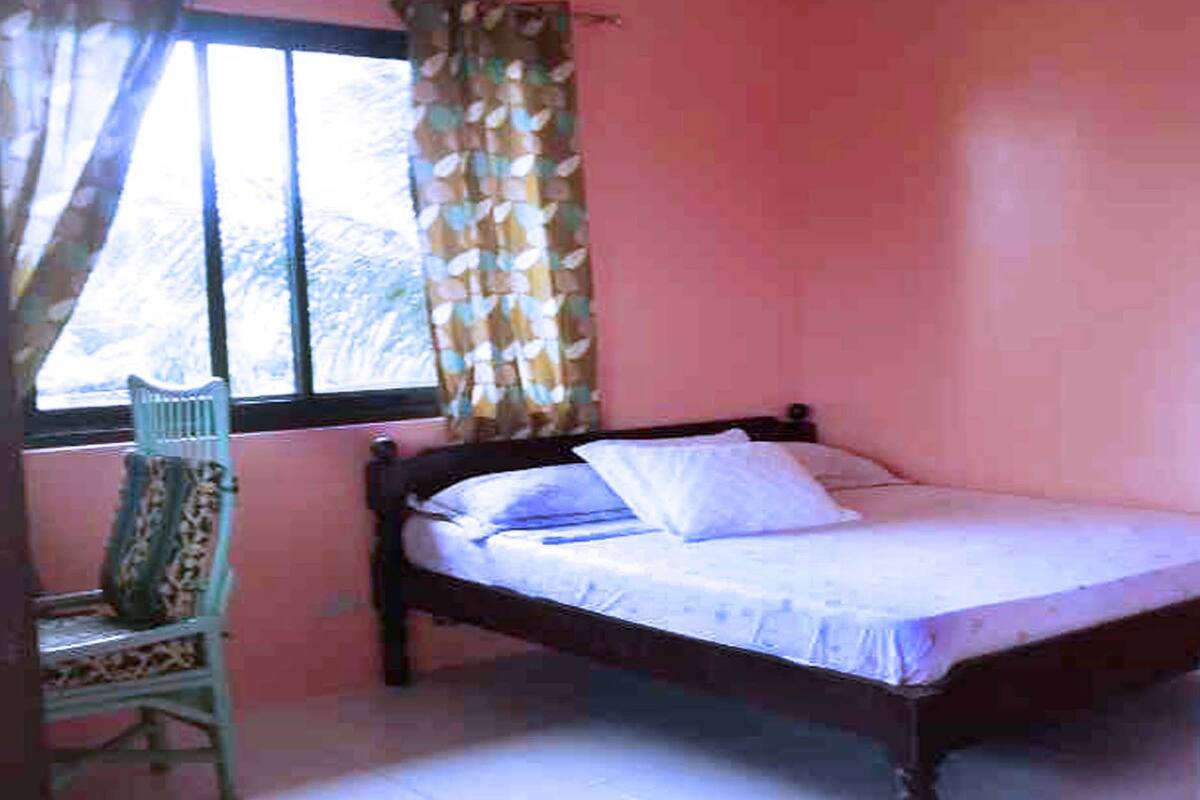 Ein Einzelbett mit weißer Matratze und Kissen, ein Holz-Kopfschild, ein Sessel mit Blumenmuster und ein Fenster mit Vorhang.