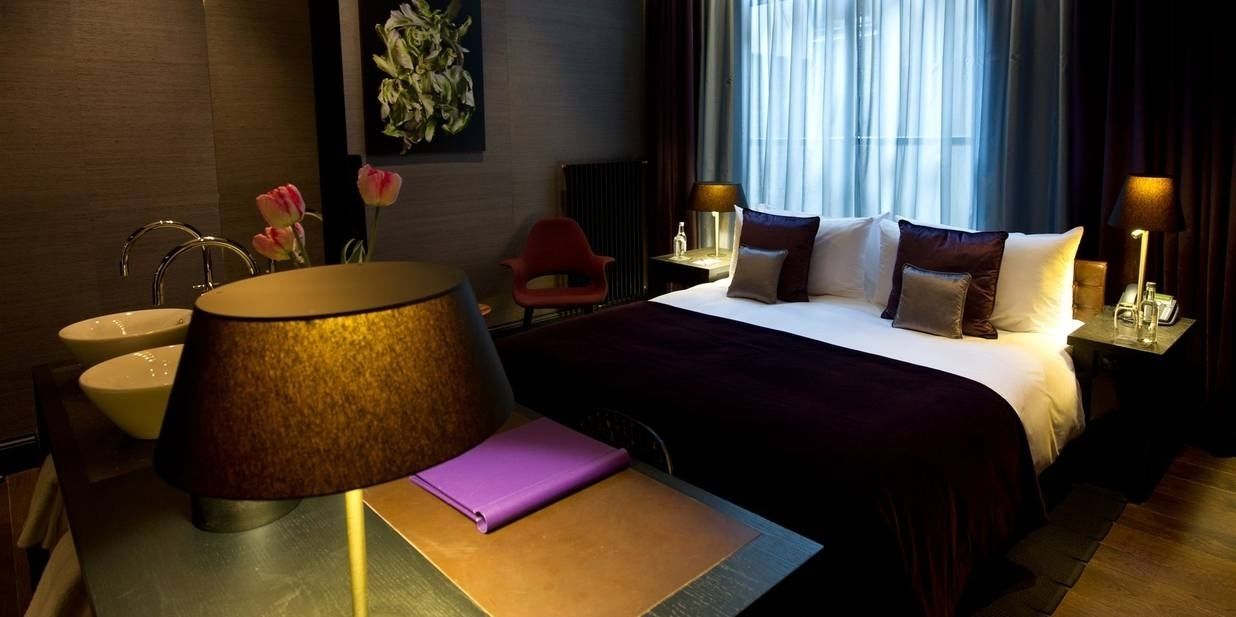 A hotel room with a bed, bedside tables, a lamp, a chair, a desk with a notebook, and a window with curtains.
