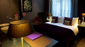 A hotel room with a bed, bedside tables, a lamp, a chair, a desk with a notebook, and a window with curtains.
