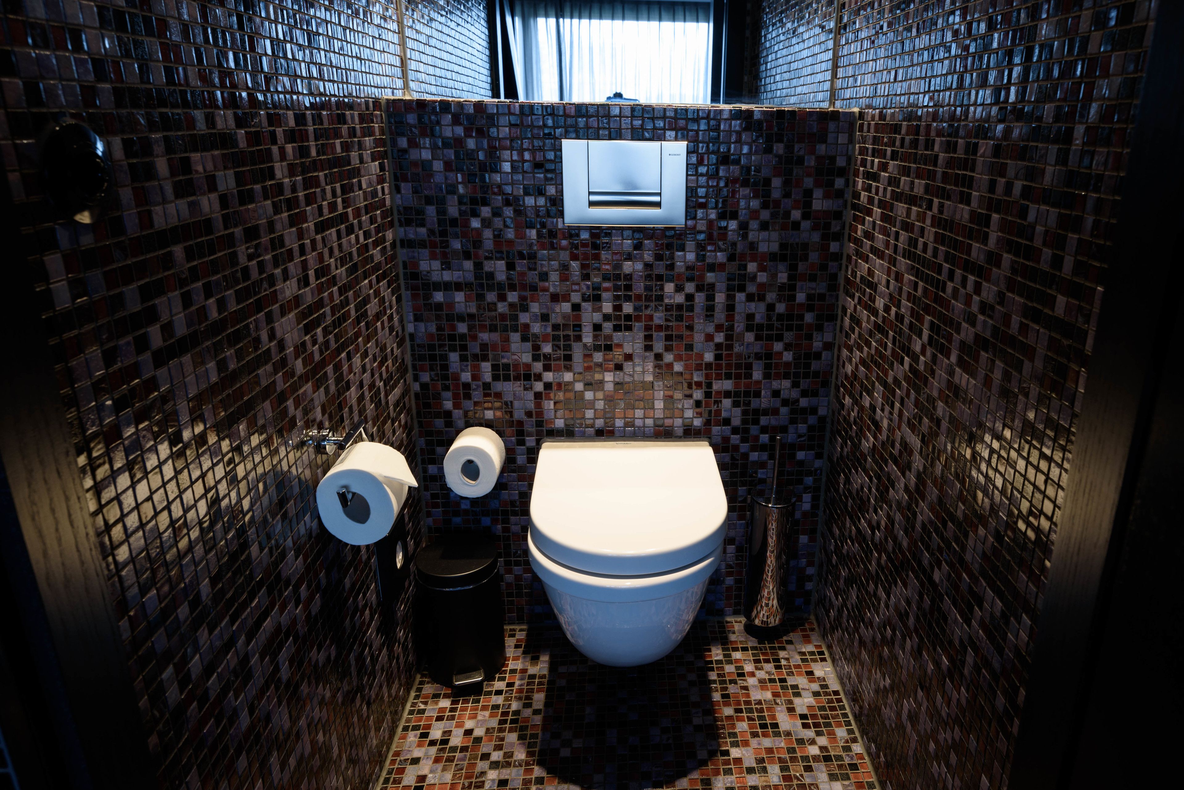 A bathroom with a mosaic tile wall, a toilet, a trash bin, and a paper towel dispenser.