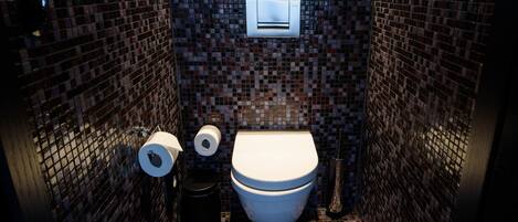 A bathroom with a mosaic tile wall, a toilet, a trash bin, and a paper towel dispenser.