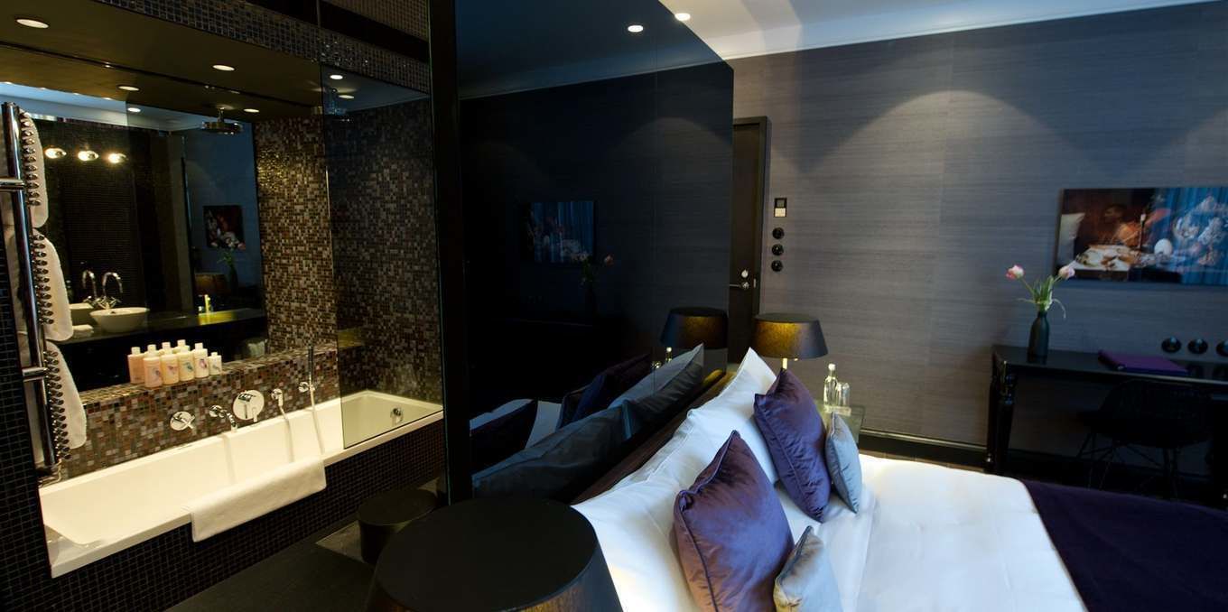 Une chambre d’hôtel moderne dotée d’un grand lit, d’un coin salon avec un bureau, et d’une salle de bain séparée avec un grand miroir et une baignoire.