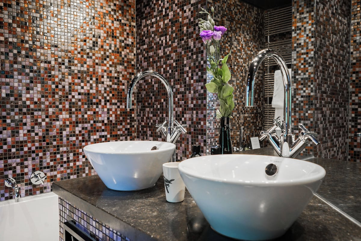 Casa de banho com duas pias, um espelho e parede revestida a mosaico.
