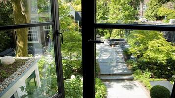 Blick durch ein Fenster auf einen Garten mit einem Weg, Sitzgelegenheiten und einem Baum.