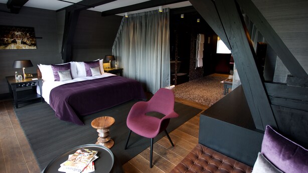 A modern hotel room with a large bed, a purple chair, a small round table, and a view of the outside. - Canal House (Amsterdam)