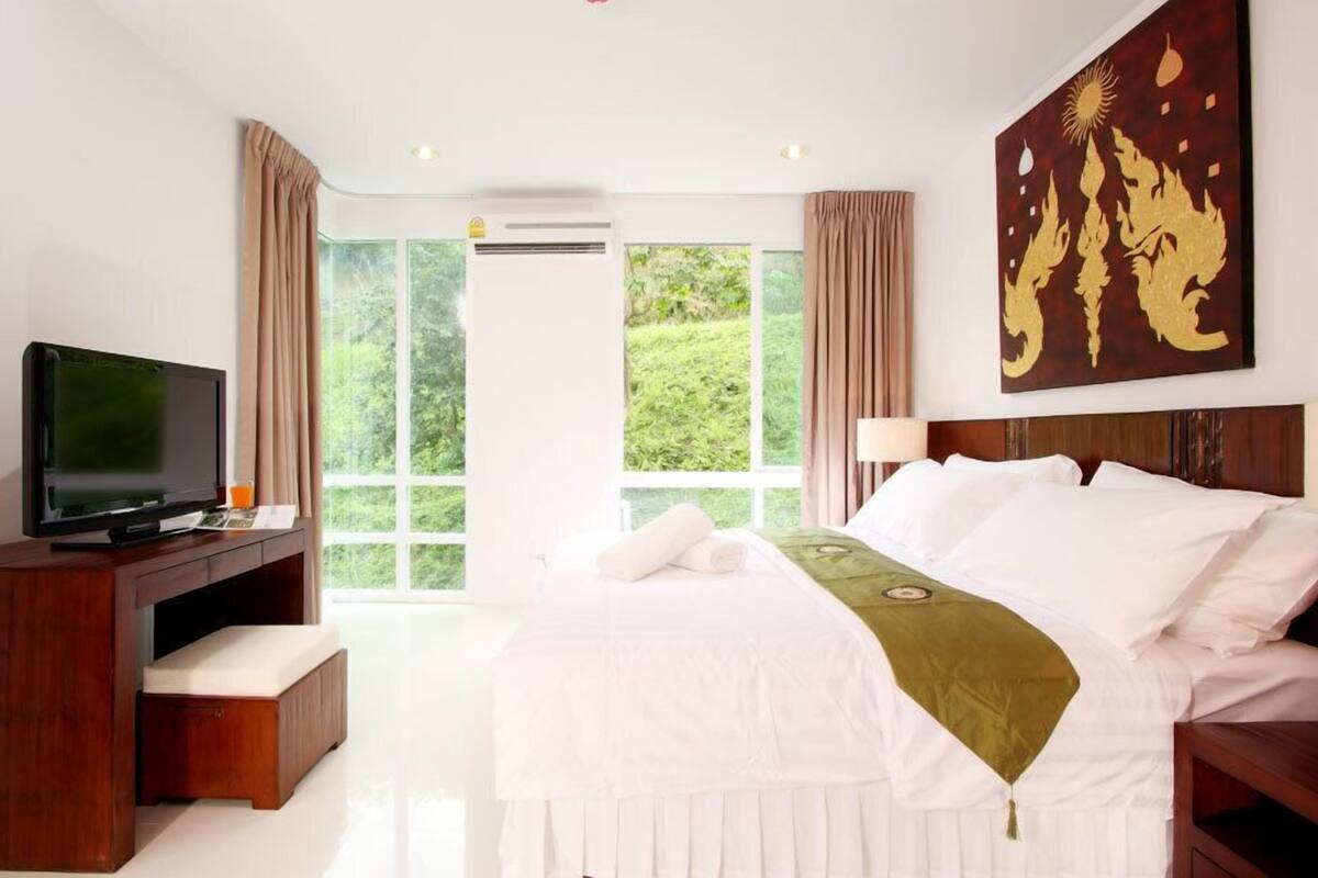 A hotel room with a bed, a TV, a desk, and a large window with curtains.