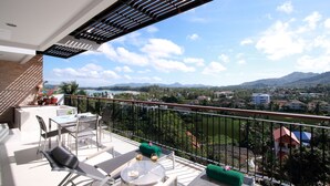 A balcony with a table and chairs, a green cushion, and a view of a city and mountains. - The Park Surin by RESAVA (Choeng Thale)