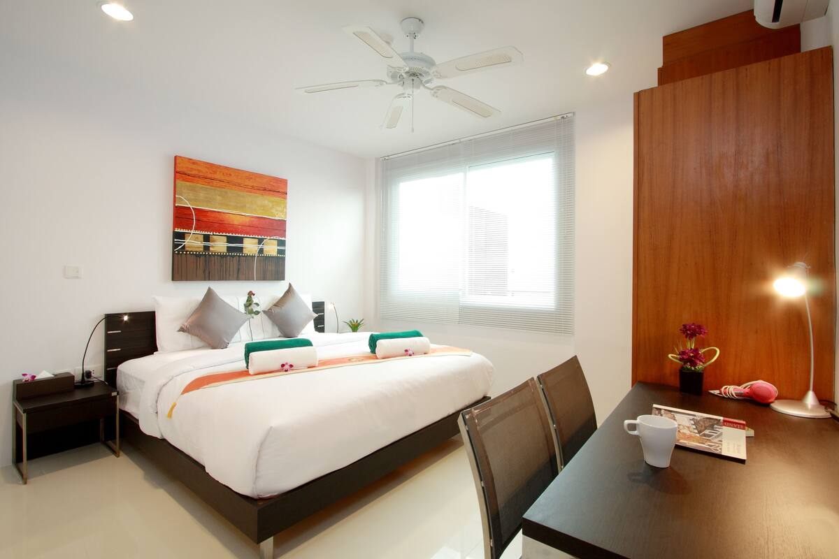 A hotel room with a bed, bedside table, chair, and a desk with a lamp and a book.