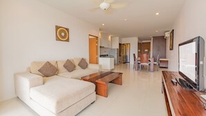 A modern living room with a sofa, coffee table, and TV. A kitchen is visible in the background. - The Park Surin by RESAVA (Choeng Thale)
