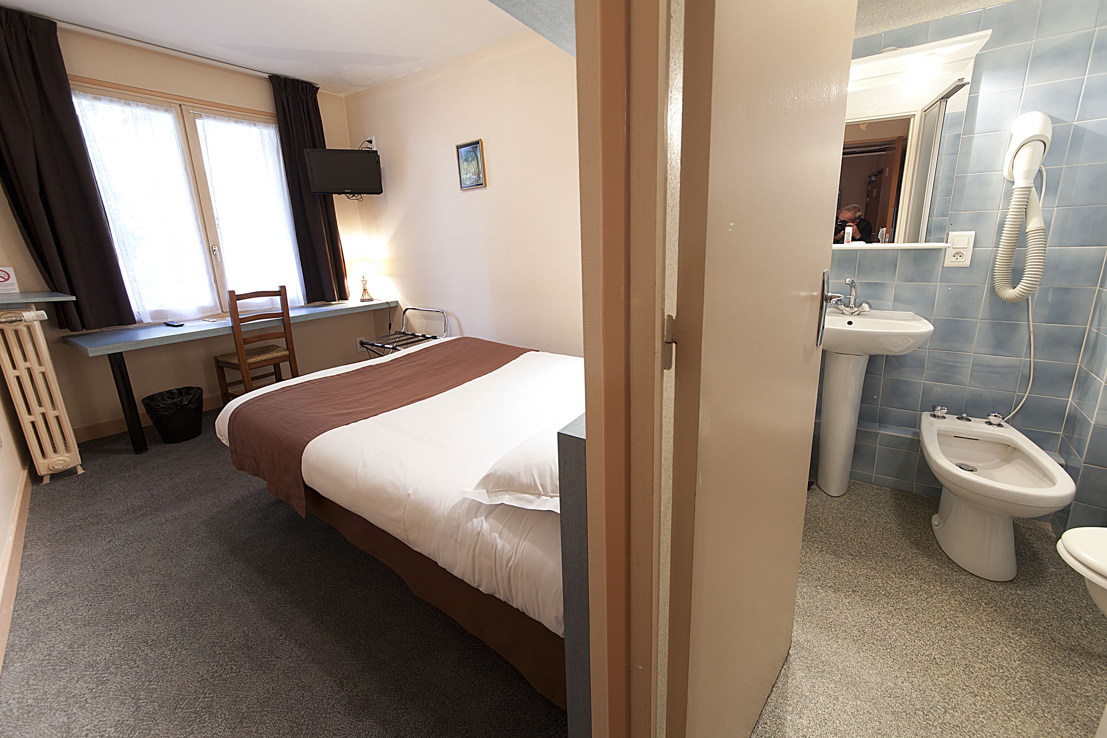 Una habitación de hotel con cama, escritorio, silla, radiador, cesto de basura, baño con lavamanos, bidé y espejo.