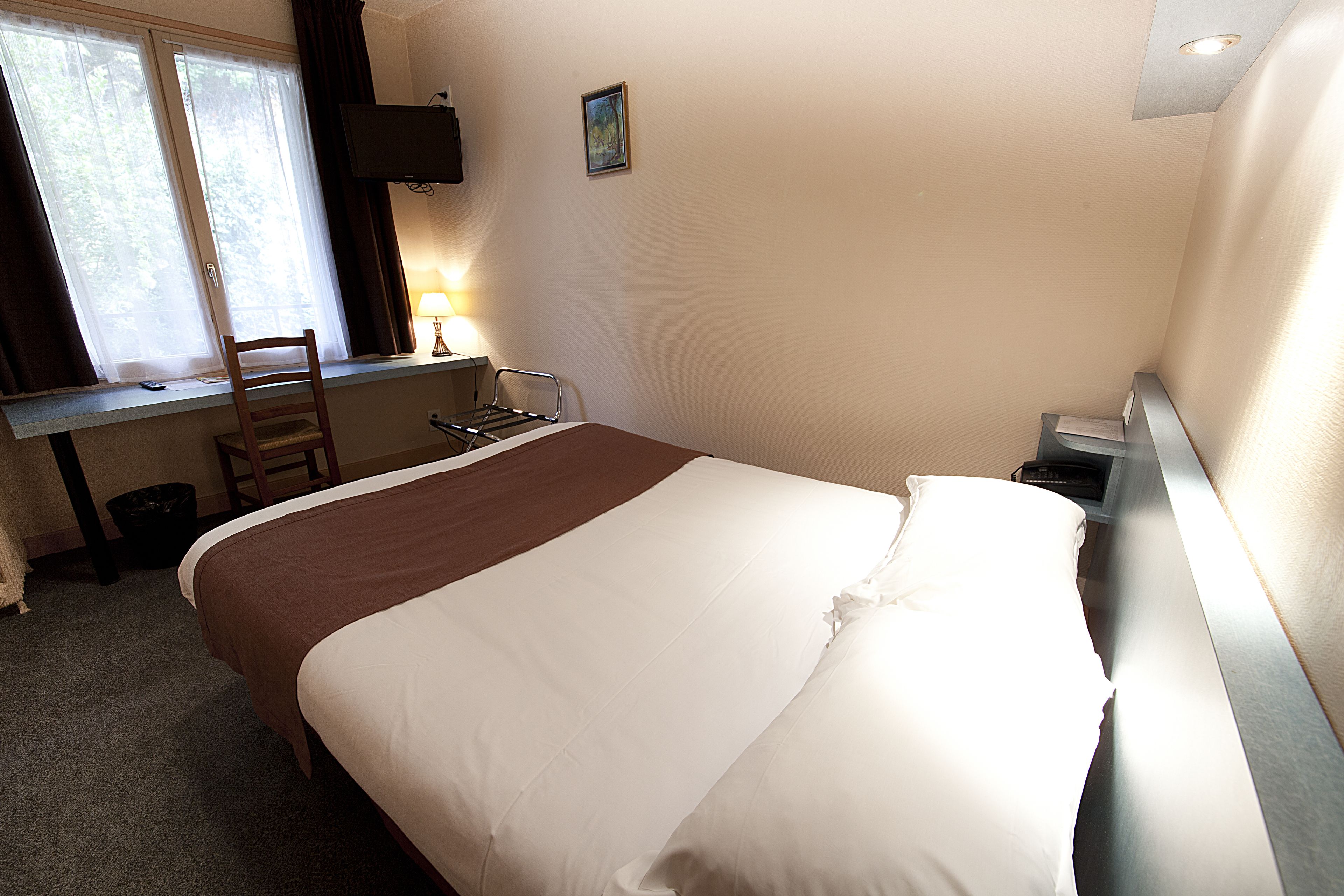 Una cama bien tendida con edredón marrón y blanco, un escritorio con silla, una lámpara y un televisor en la pared.