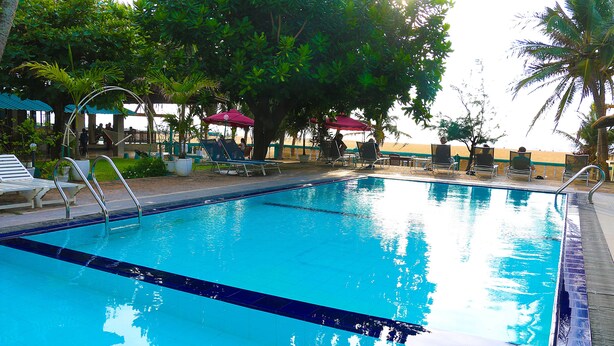 A clear blue swimming pool with lounge chairs and umbrellas, surrounded by trees and a view of the beach. - Topaz Beach Hotel (Negombo)