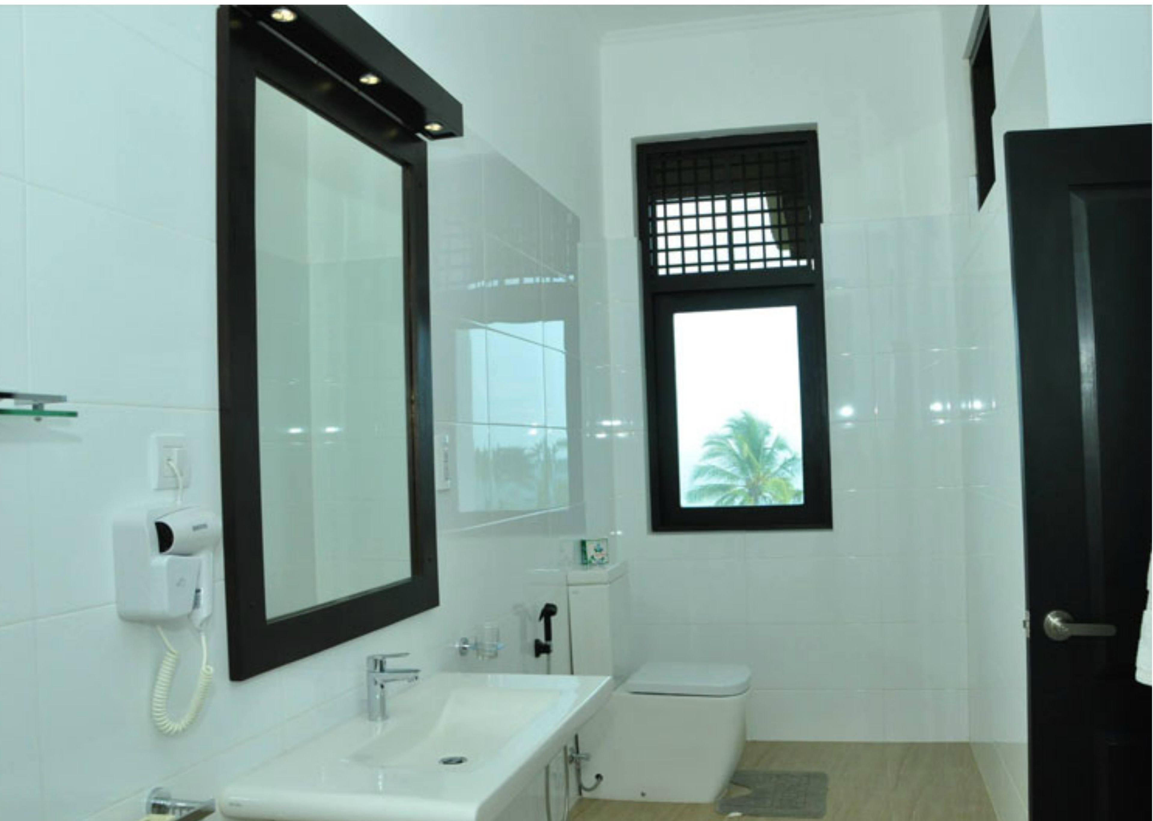 A modern bathroom with a large mirror, a sink, a toilet, and a window with a view of palm trees.