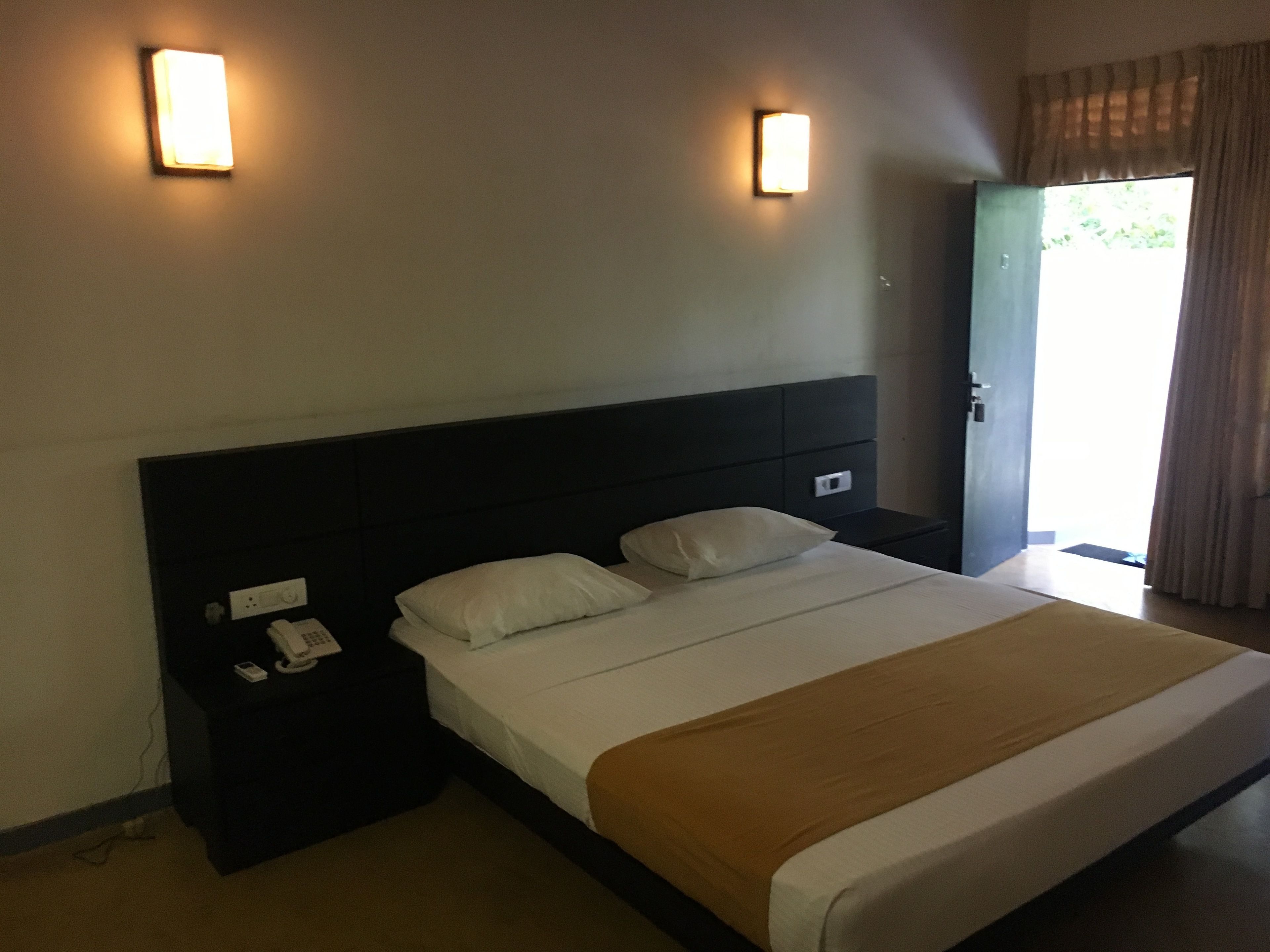 Habitación de hotel con una cama grande, dos mesitas de noche, un teléfono sobre una de ellas y una puerta que da a un balcón.
