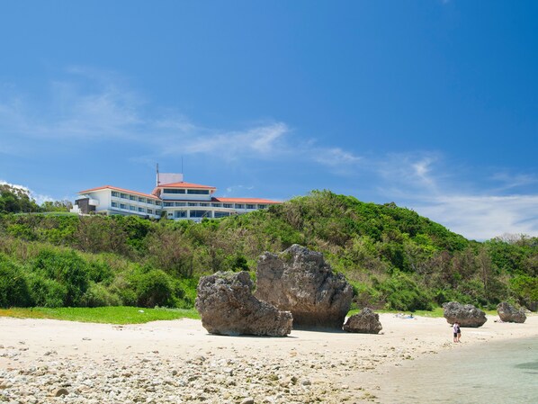 A beachfront hotel with a large building, rocky beach, and clear skies. - Hotel Hamahigashima Resort (Uruma)