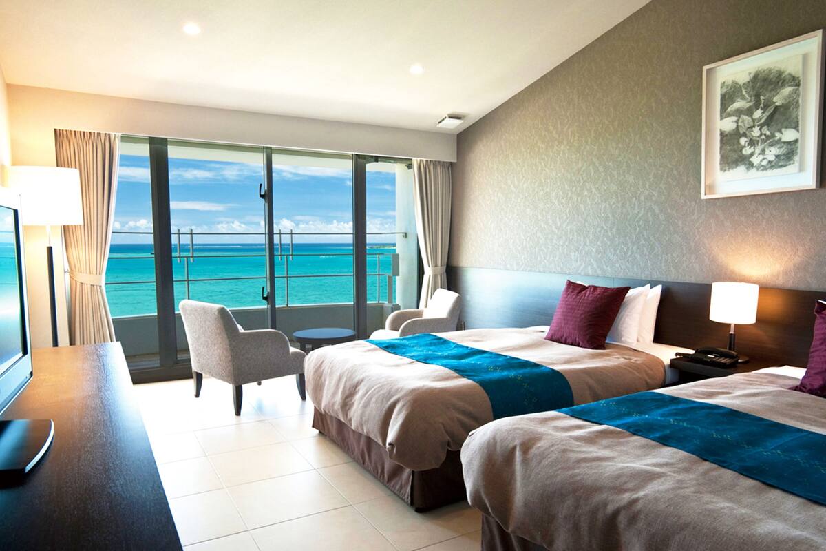 Habitación de hotel con dos camas, un escritorio con silla, televisor y vista al océano.