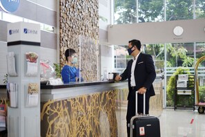 A hotel reception desk with a modern design, a clock on the wall, and a view of greenery outside.