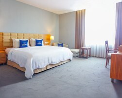 A hotel room with a large bed, a nightstand, a desk, a chair, and a view of the outdoors.