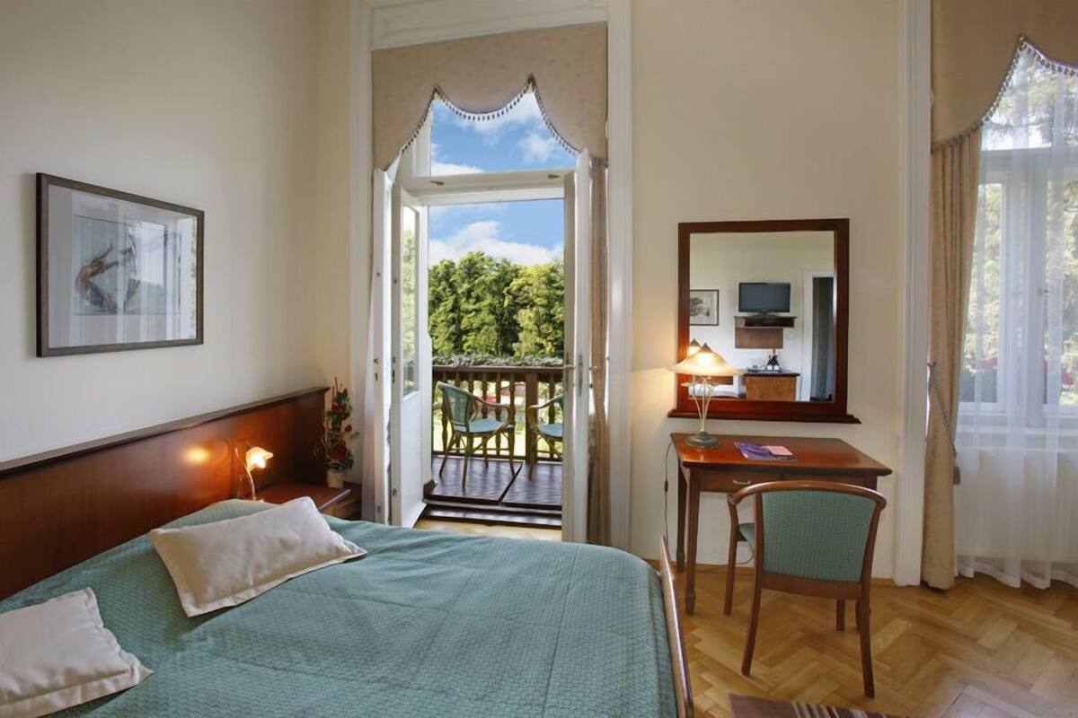 Habitación de hotel con cama, escritorio y silla, televisor y balcón con sillas.