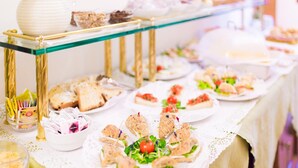 Una mesa de buffet con diversos alimentos, como sándwiches y ensaladas, dispuesta sobre una mesa de vidrio y dorado con mantel de encaje blanco.