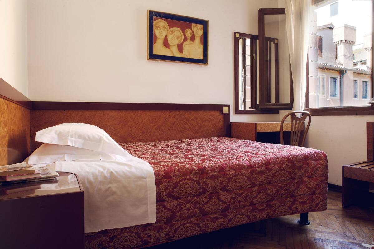 A hotel room with a wooden headboard, a bed with a red patterned bedspread, a white pillow, a wooden nightstand with books, a framed painting on the wall, and a window with a view of buildings.