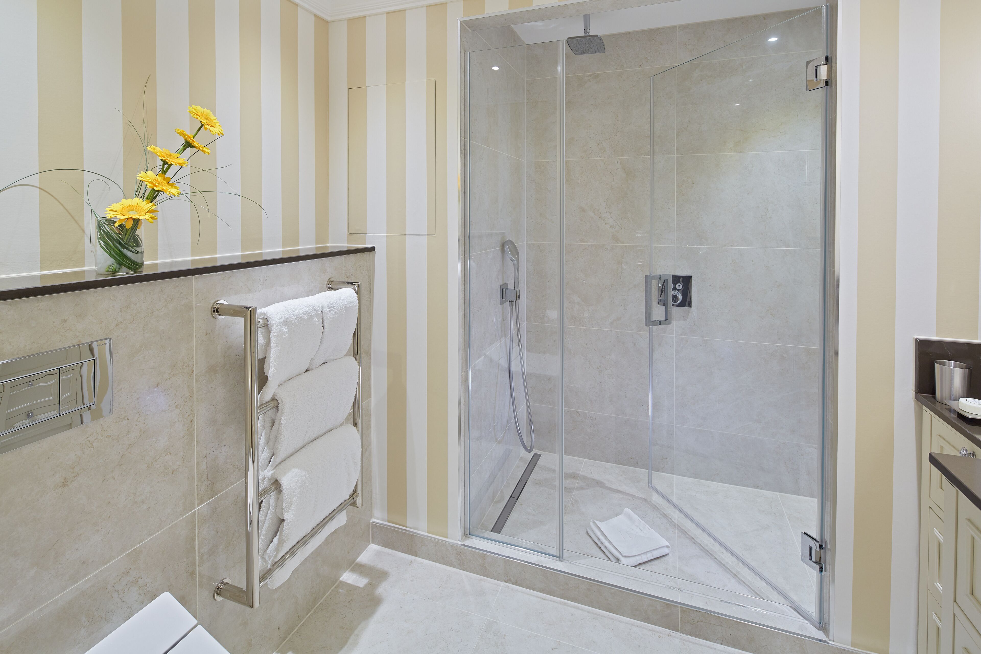 A modern bathroom with a glass-enclosed shower, a towel rack with folded towels, and a vase with yellow flowers on the countertop.